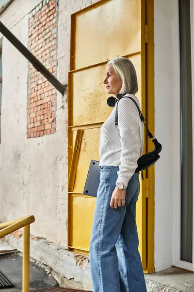 A stylish woman with gray hair awaits by a bright yellow door, showcasing elegance and confidence. — Stock Photo