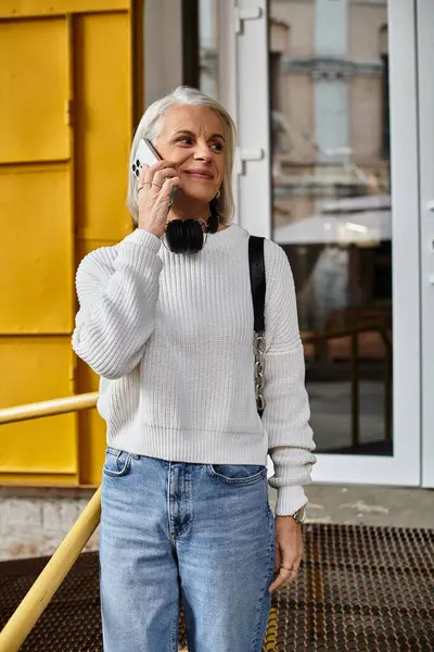 A mature woman in a cozy sweater chats on her phone by a vibrant yellow backdrop. — Stock Photo