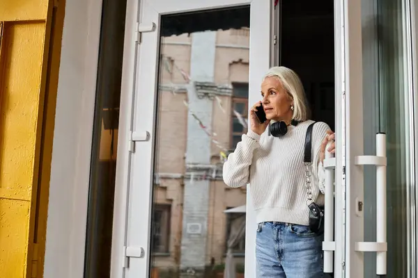 Una mujer madura en un suéter acogedor se encuentra en la puerta, comprometida en una conversación telefónica. - foto de stock