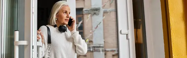 A stylish older woman is talking on her phone while stepping through an open door. — Stock Photo