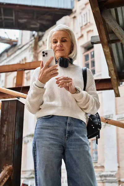 A stylish mature woman with gray hair enjoys a moment on her phone outside in the city. — Stockfoto