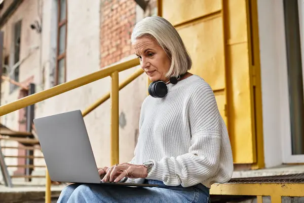 A mature woman with gray hair sits on steps, focused on her laptop and wearing headphones. — Stock Photo