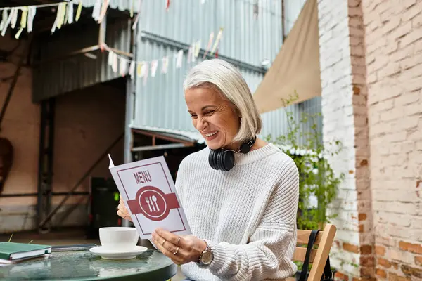 A joyful woman with gray hair sits at a cafe table, eagerly reading the menu while sipping coffee. — Stock Photo