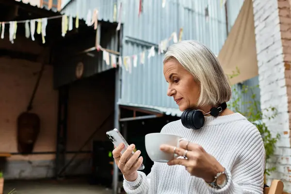 A mature woman with gray hair sits comfortably, sipping coffee and texting on her phone. — Stock Photo