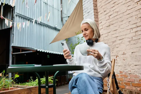 A beautiful woman with gray hair relaxes at a cafe, sipping coffee and using her smartphone. — Stock Photo