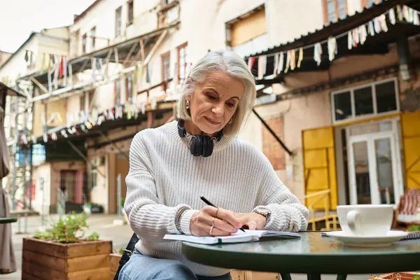 Die schöne grauhaarige Frau sitzt an einem Tisch und schreibt aufmerksam in einem belebten Hof. — Stockfoto