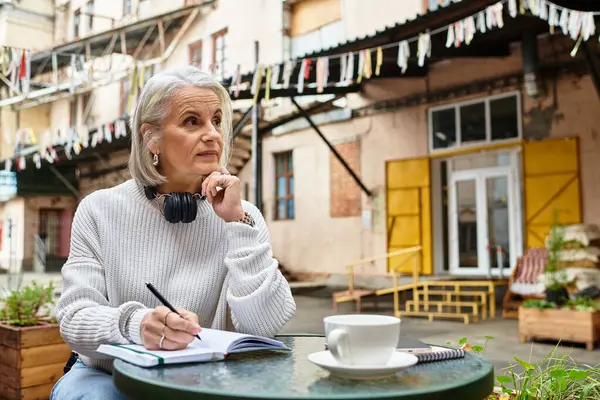 A thoughtful mature woman sits at a table enjoying coffee and writing in a notebook. — Stock Photo