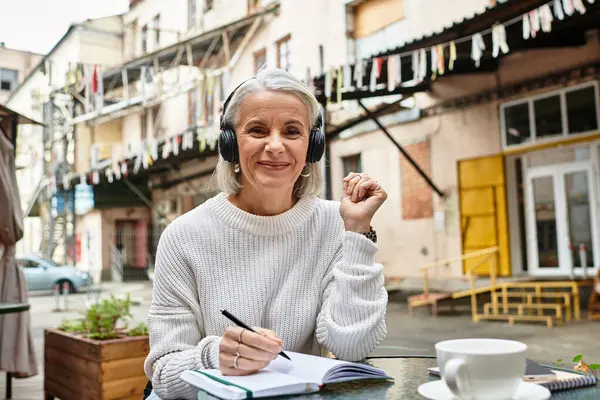 Una mujer sonriente con el pelo gris escribe en su cuaderno mientras toma café en un animado café al aire libre. — Stock Photo