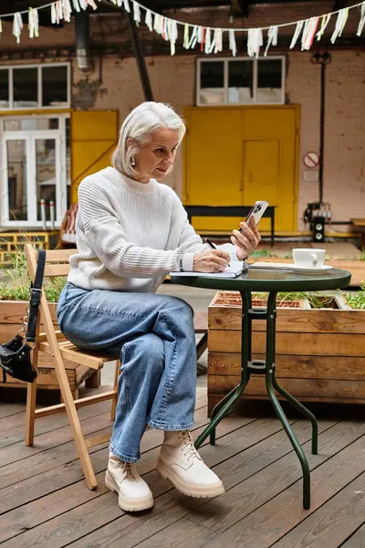 Una donna matura con i capelli grigi siede a un tavolo da caffè, scrivendo note mentre si gode una bevanda calda. — Foto stock