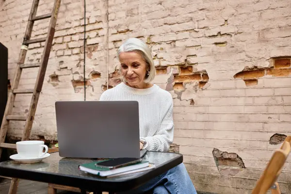A lovely woman with gray hair sits at a table, immersed in her laptop, sipping coffee. — Stock Photo