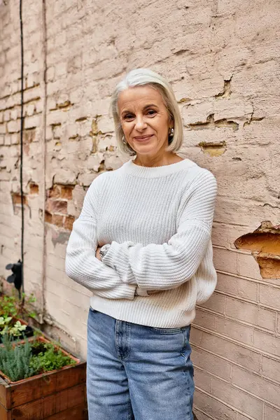 A graceful mature woman with silver hair enjoys a serene moment against a charming brick wall. — Stock Photo
