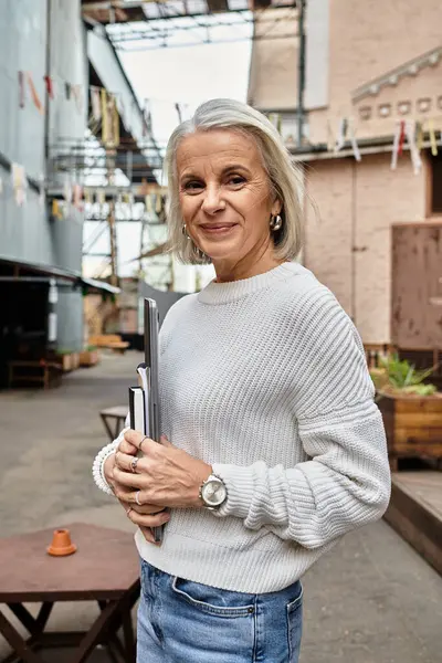Una mujer confiada con el pelo gris muestra su estilo mientras sostiene un cuaderno en un espacio urbano. — Stock Photo