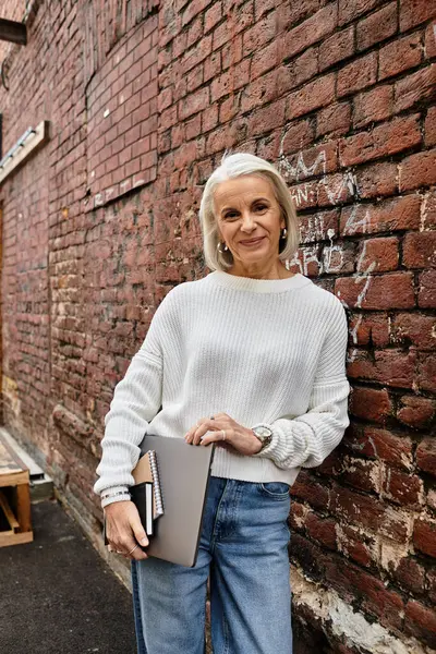A graceful woman with gray hair holds a notebook by a textured brick wall. — Stock Photo