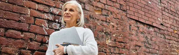 Uma mulher madura com belo cabelo grisalho fica confiante com um caderno, sorrindo suavemente. — Fotografia de Stock
