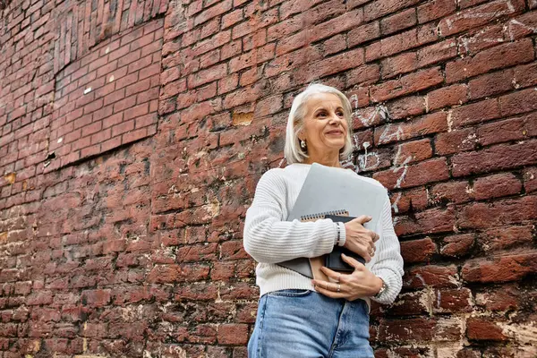 A mature woman with gray hair embraces a notebook while standing outdoors, smiling gently. — Stock Photo