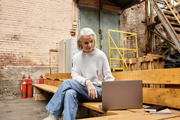 A beautiful mature woman with gray hair focuses on her laptop while seated in a creative space. — Stock Photo