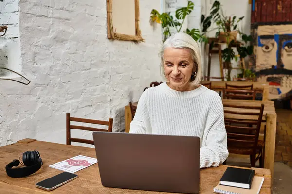 A mature beautiful woman with gray hair engaged in focused work on her laptop in a vibrant cafe. — Stock Photo
