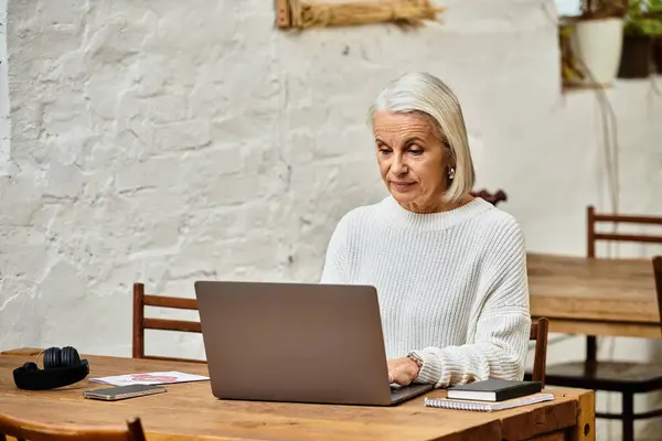 A beautiful mature woman is focused on her laptop while seated at a wooden table in a cafe. — Stock Photo