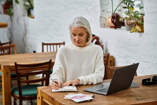 A mature woman with gray hair sits at a wooden table, focused on writing in her notebook. — Stock Photo