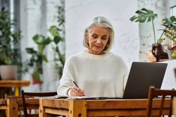 A beautiful mature woman with gray hair writes intently at a wooden table in a sunny cafe. — Stock Photo