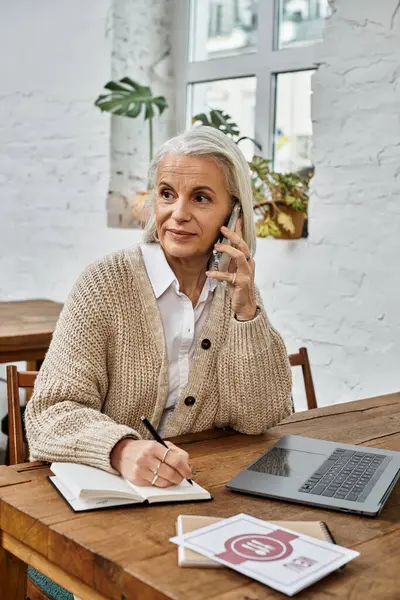 A sophisticated woman with gray hair takes a call, jotting down important notes in a cozy setting. — Stock Photo