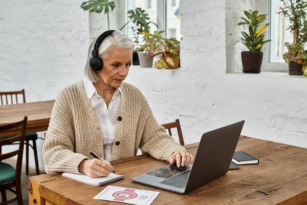 A mature woman with silver hair engages in an online session while seated at a wooden table. — Stock Photo
