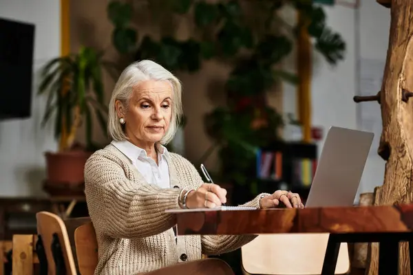 A beautiful woman with gray hair diligently writes in a notebook while sitting at a wooden table. — Stock Photo