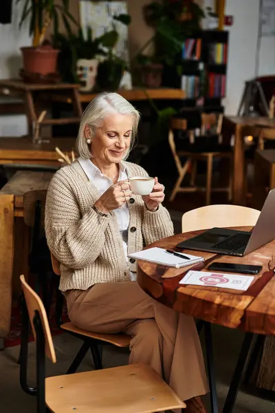 A beautiful mature woman with gray hair savors a drink, immersed in her work at a cafe. — Stock Photo