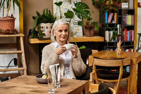 In a charming cafe, a mature woman with silver hair savors her tea while relaxing at a wooden table. — Stock Photo