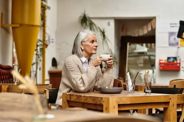 A beautiful mature woman with gray hair sits thoughtfully in a warm cafe, sipping her drink. — Stock Photo