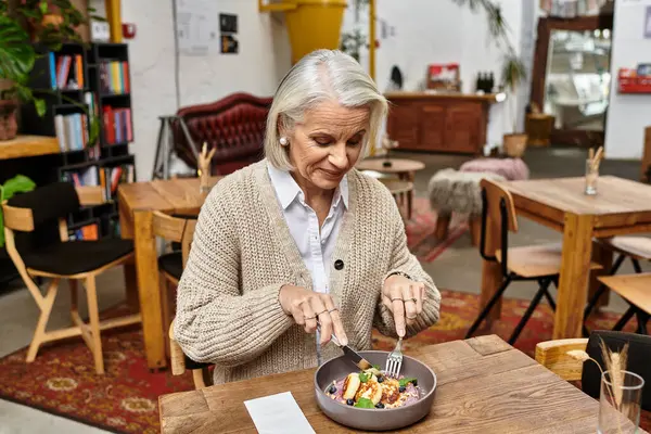The mature woman with gray hair savors a colorful dish while seated at a wooden table. — Stock Photo