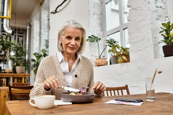 A smiling woman with gray hair savors her food in a bright, plant filled cafe. — Stock Photo
