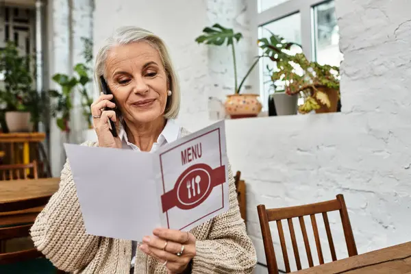 A sophisticated woman with gray hair reads the menu and chats on her phone in a cozy cafe. — Stock Photo