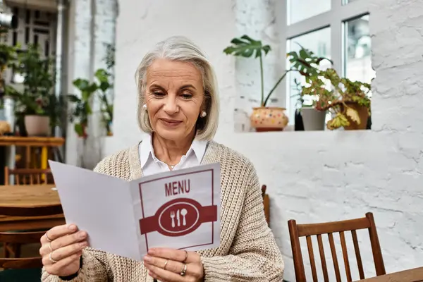 A mature woman with gray hair browses the menu at a cozy, plant filled cafe. — Stock Photo