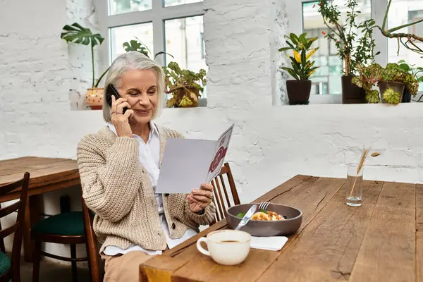 Uma bela mulher de cabelos grisalhos senta-se em uma mesa de madeira, desfrutando de café da manhã e fazendo uma chamada. — Fotografia de Stock