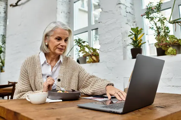 A graceful gray haired woman sits at a table, enjoying her meal while using her laptop. — Foto stock