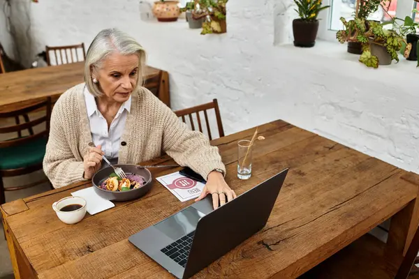 A mature woman with gray hair savoring a healthy dish while engaged with her laptop at a cozy cafe. — Stock Photo