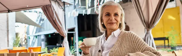 A charming woman with gray hair sits relaxed in a cafe, sipping tea and enjoying the ambiance. — Stockfoto