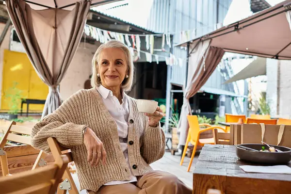 This elegant woman with gray hair relaxes at a cafe, savoring her tea in a charming setting. — Stock Photo