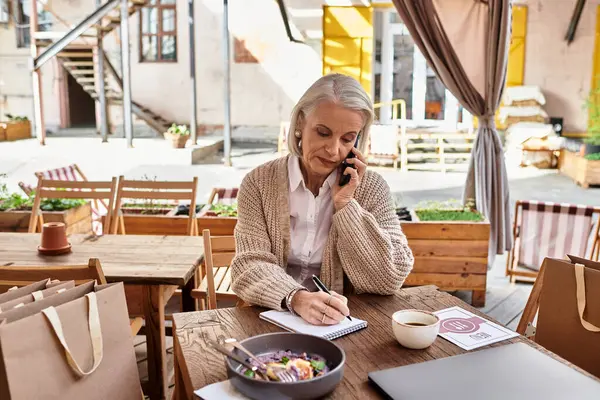 A mature woman writes in her notebook while talking on the phone at a charming outdoor cafe. — Stock Photo