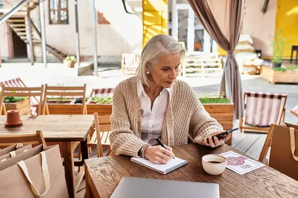 A mature woman with gray hair sits at a wooden table, sipping coffee and writing in her notebook. — Stock Photo