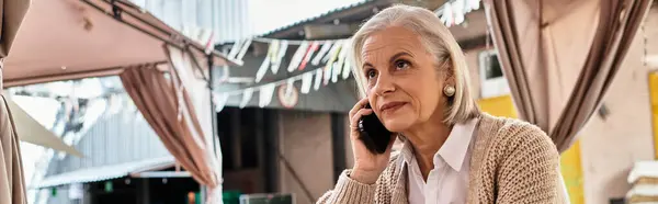 Una bella donna anziana con i capelli grigi parla al telefono sotto un baldacchino colorato. — Foto stock