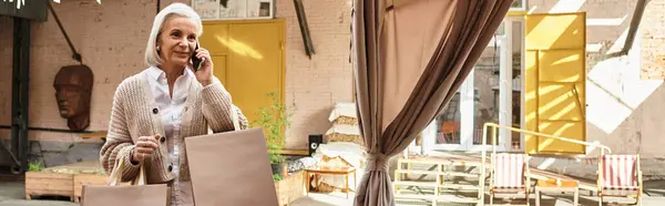 A beautiful older woman carries shopping bags while engaged in a phone call outdoors. — Stock Photo