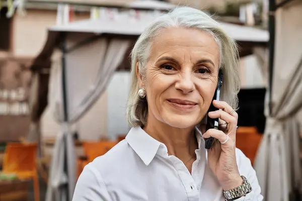 A graceful woman with gray hair smiles as she talks on her phone in a lovely outdoor setting. — Stock Photo