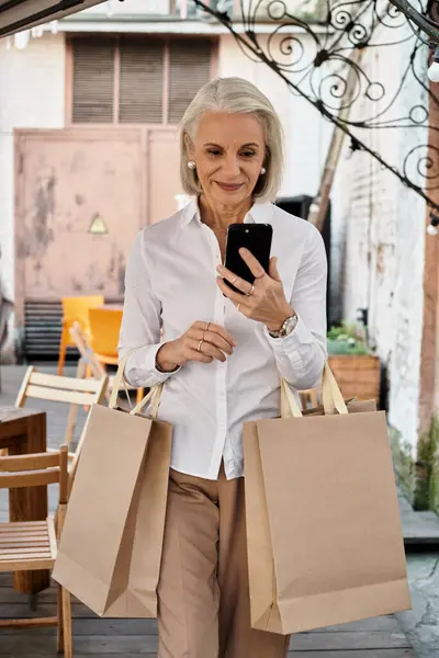 Una donna anziana elegante gode la sua giornata fuori, sorridendo mentre guarda il suo telefono, tenendo borse della spesa. — Foto stock