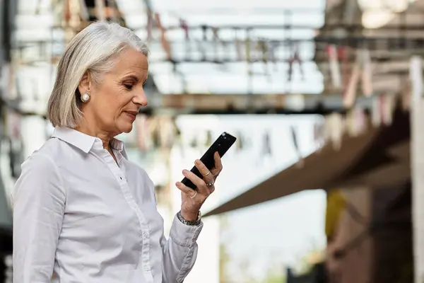 A mature woman admires her smartphone in a vibrant outdoor market filled with colors. — Stock Photo