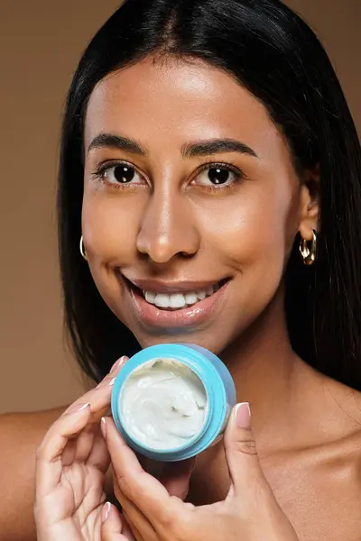 Una mujer muestra su rutina de cuidado de la piel mientras sonríe y sostiene un recipiente de crema en un estudio. - foto de stock