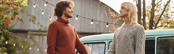 Jeune couple gay amoureux vêtu de pulls chauds partager un moment dans un paysage d'automne. La douce lueur des lumières à cordes illumine leur environnement, créant une atmosphère chaleureuse près d'un van vintage. — Photo de stock