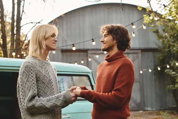 Two young men share a warm gaze while holding hands, surrounded by softly lit autumn ambiance. The backdrop features a vintage green van and trees, creating a romantic atmosphere. — Stock Photo