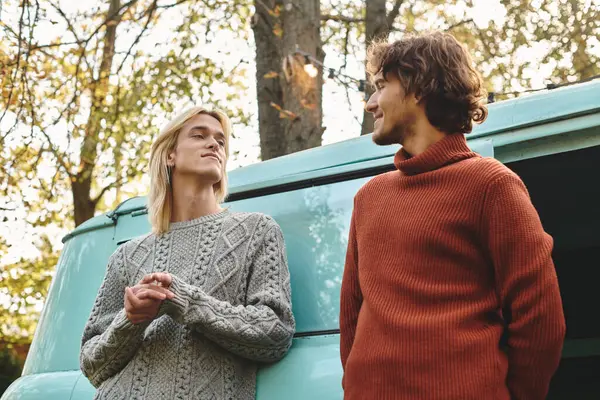 Two stylish young men share a warm moment outdoors during autumn, standing beside a vintage camper van. The soft light highlights their happiness amid the colorful leaves. — Stock Photo